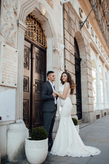 Young romantic couple standing and hugging at city street on a sunny summer day. Handsome guy in suit and girl in white dress with a train. A love story. Beautiful young man and woman pose
