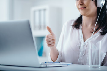 Woman with headphones at the workplace. Giving thumbs up at the screen