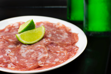 Sliced salami in a plate close in black background with two green bottles of beer in the background