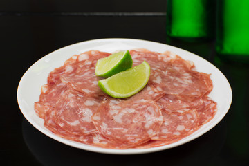 Sliced salami in a plate in black background with two green bottles of beer in the background