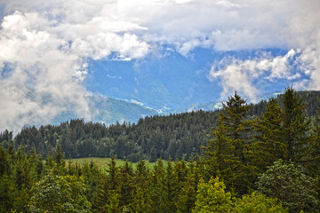 La Clusaz, France - August 9th 2017 :  View on a valley, with fir forests, grass field and cloudy big blue mountains in background.