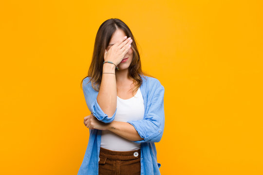 young pretty woman looking stressed, ashamed or upset, with a headache, covering face with hand against orange wall