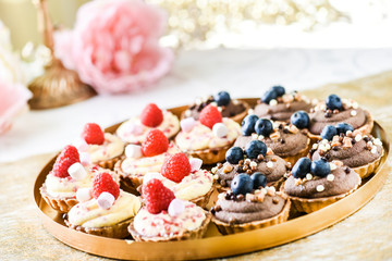 Small cakes with chocolate caramel and blueberries, raspberries on top. Finest cupcakes desserts on gold tray.