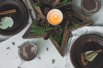 Festive Christmas and New Year table setting in scandinavian style with rustic handmade details in natural and white tones. Dining place decorated with handmade dishes, pine cones,branches, candles an