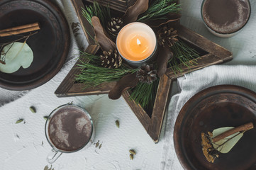 Festive Christmas and New Year table setting in scandinavian style with rustic handmade details in natural and white tones. Dining place decorated with handmade dishes, pine cones,branches, candles an