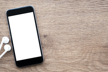 Black smartphone with blank mock up screen is on top of wood office desk table with earphones. Top view with copy space, flat lay, close up to the phone.