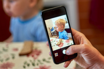 Hand holds smartphone photographing little boy sitting at the table