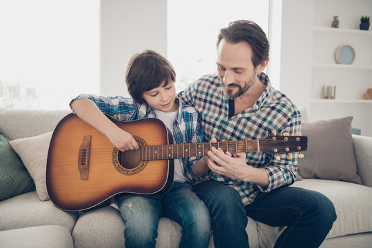 Best weekend activities ever concept. Photo of satisfied mature bearded daddy enjoying her son playing guitar sitting on comfortable sofa wearing checkered plaid shirt singing melody
