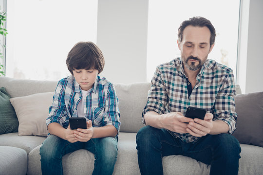 Do not leave in virtual life spend time with your relatives concept. Photo of serious kid and his bearded in checkered shirt dad holding telephones in hands typing request on internet watch video