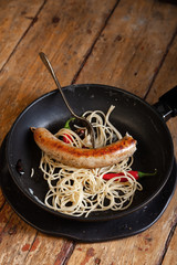 Hearty rustic bachelor's breakfast Italian pasta spaghetti, bitten off fried meat sausage, red chili pepper in black frying pan, fork. Still life. Closeup, wooden background