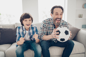 Portrait of two nice attractive lovely cute cheerful cheery overjoyed guys dad and pre-teen son sitting on sofa watching sport show online in light white modern style interior house living-room