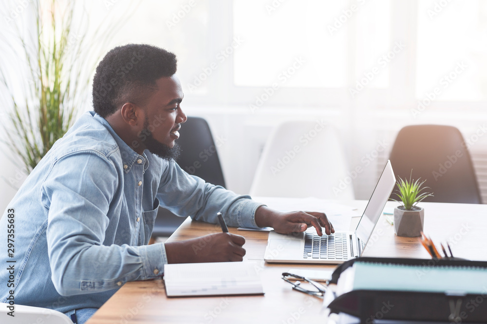 Poster african worker taking notes while using laptop in office