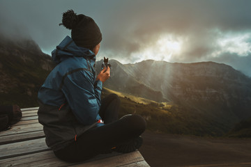A hiker girl sits on the viewpoint and eats an energy bar, watching the sunset on the mountains