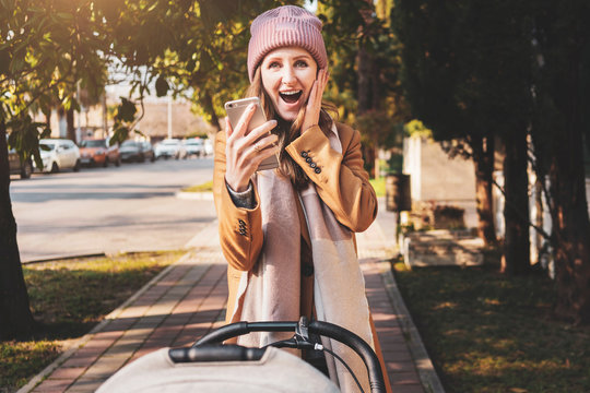 Emotional young woman walking with baby stroller along street and shooting stories on mobile phone. Young mother taking selfie with emotions on her face. Girl watching fantastic content on smartphone.