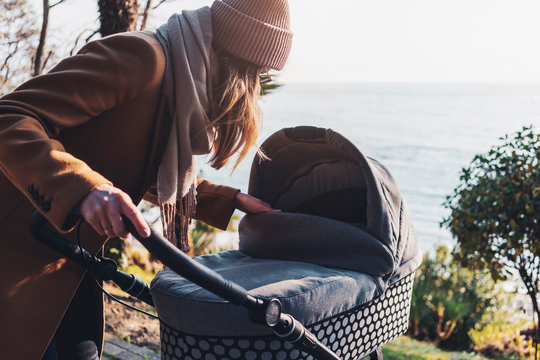 Young Mother With Baby Stroller Walking By Seaside. Woman Caring About Child, Walking Outdoors Down Coastline. Casual Style For Moms With Little Children. Trees, Bushes, Warm Weather In Autumn, Fall.