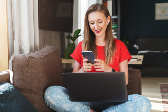 Young woman wearing casual clothes sitting indoors at home and texting on smartphone. Girl smiling and looking at mobile device screen, listening to music in earphones, watching videos, making calls.