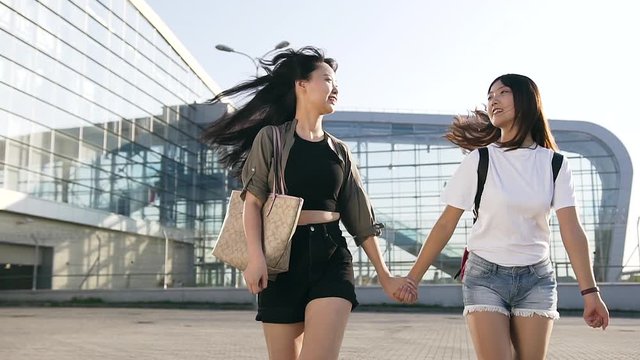 Attractive picture of happy smiling young asian girls in trendy clothes holding by hands and having fun together near the airport building in day time