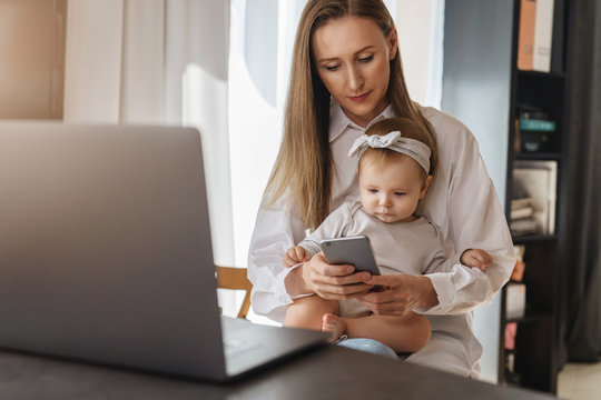 Young woman texting on smartphone with baby girl sitting on her knees and looking at screen. Mother working at home distantly using wireless connected devices for job. Freelance vacancies for parents