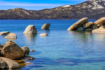 Clear beautiful blue lake with boulders in foreground, mountains with snow in background.