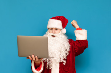 Portrait of joyful man in santa costume stands against blue background and rejoices with raised hand, looks into screen. Happy young Santa rejoices at victory with laptop in hands.