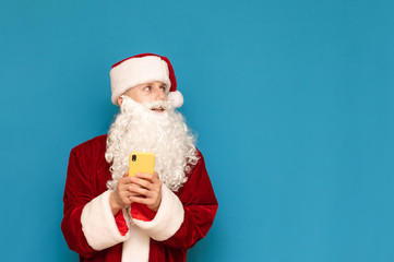 Cheerful young man Santa Claus stands with a smartphone in his hands, looks away at an empty space and smiles. Positive guy in santa suit looking away at copy space, holds phone in his hand