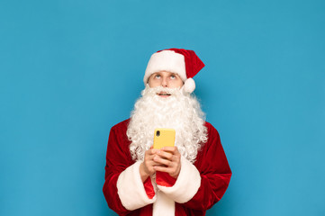 Funny young santa stands on blue background with smartphone in hands and looks up at empty space. Cheerful Santa is using a smartphone and looking up at copyspace. New Year and Christmas concept.