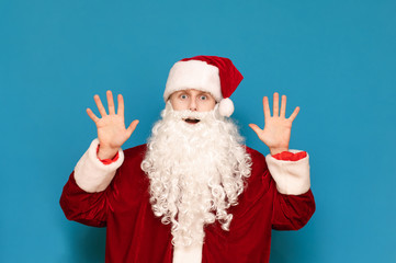 Fototapeta na wymiar Portrait of a frightened young Santa on a blue background, raised from shocked hands up and looks into the camera with an emotional face. Shocked guy in santa costume isolated. Christmas concept.