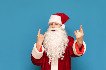 Cheerful young Santa Claus shows heavy metal sign and looks into camera, guy in santa costume loves to listen to rock music. Playful Santa shows rock gesture and poses on camera.