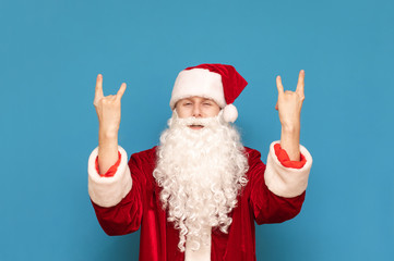 Young Santa Claus rocker stands on a blue background and shows with his hands a sign of Heavy Metal looking into the camera.Santa listens to rock music, shows rock sign.Portrait of Rock and Roll Santa