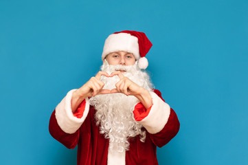Handsome young Santa Claus shows heart gesture with his hands and looks into the camera with a positive face isolated on a blue background. Santa loves you. Christmas and New Year.