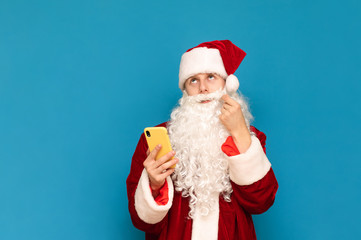Portrait of a pensive Santa Claus using a smartphone, twirling his mustache and looking thoughtfully upwards against a blue background. Guy in santa suit and smartphone in hand is isolated. New Year