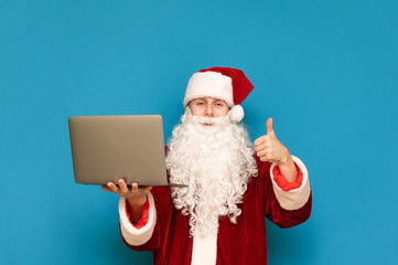 Cheerful young man in santa costume holds laptop in hand and shows thumb up on blue background. Happy Santa Claus freelancer with laptop isolated. Copy space