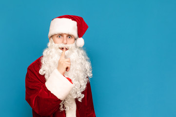 Scared boy in santa costume looks into camera with shocked face and shows gesture of silence with hand on blue background. Young Santa asks for silence, isolated. Tshshsh. Copy space