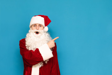 Portrait of surprised young Santa Claus, isolated on blue background, pointing finger at copyspace, looking into camera with surprised face. Shocked young Santa is showing up to empty space