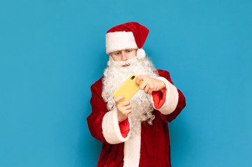 Guy in carnival costume of santa claus playing mobile games on smarphone isolated on blue background. The young site plays online games and smartphones, looks intently at the smartphone screen.
