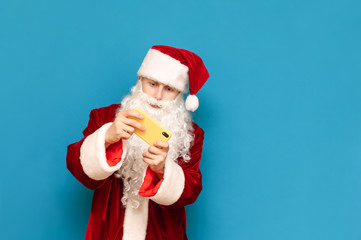 Focus on the phone. Concentrated guy in Santa costume playing mobile games on smartphone, looking intently at the screen. Young Santa gamer playing video games on smartphone. Christmas mobile gaming.
