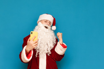 Young Santa Claus winner rejoices in victory with smartphone in hand on blue background, looks into camera with happy face. Successful gamer plays mobile games on smartphone. New Year and Christmas.