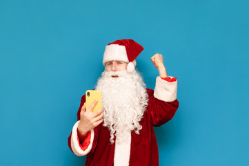 Portrait of a joyful young Santa Claus playing video games on smartphone, rejoices at winning on a blue background, looks into the camera with a happy face. Guy gamer in Santa costume is happy to win