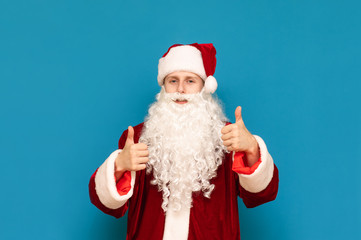 Fototapeta na wymiar Happy young Santa Claus in suit standing against blue wall background and showing thumb up, looking into camera and smiling. Santee likes it. Christmas and New Year concept.
