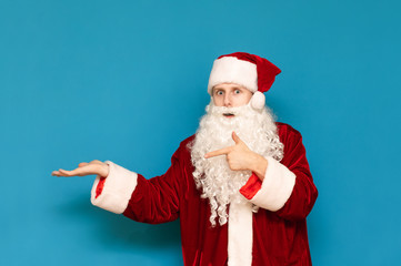 Fototapeta na wymiar Surprised young Santa Claus in hat and white beard, looks at camera and shows his hand on copy space. Isolated on blue background. Amazed Santa is pointing to an empty space. Background