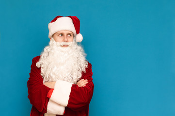 Young guy in santa costume stands on blue background and looks away at blank space, isolated. Young funny santa on blue wall background. Copyspace. Christmas concept.