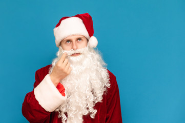 Closeup portrait of a funny santa on a background of blue wall, spinning mustache and looking up. Young guy in santa suit isolated on blue background. New Year concept.