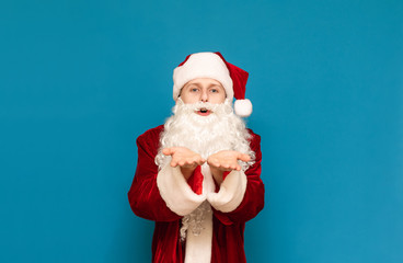 Portrait of positive young man in santa costume looking into camera and sending a kiss on a blue background. Santa holds palm near face and blows into camera.