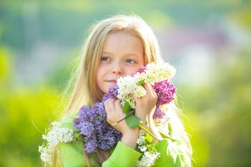 Fototapeta premium A good-looking fashionable little girl in the garden with long fair hair being excited to get a bouquet of lilac on a birthday.