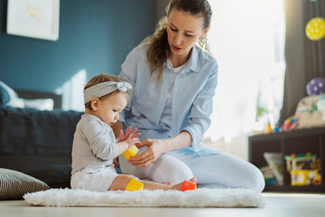 Mother sitting with baby on floor and playing toys. Educating games for infants and toddlers. Babysitting services for parents.