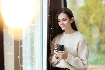 Portrait of a young beautiful woman in a warm sweater and with a cup of hot drink at home on the background of a window. Lamps of warm light.