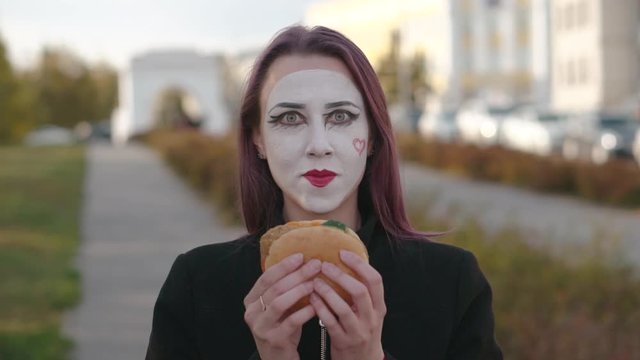Mime girl in a black coat eats burger on the street. Slow motion.