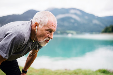 A senior man with earphones standing by lake in nature, doing exercise.