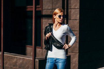 Stylish model girl at the city in black leather jacket and white sweater and glasses with waist bag