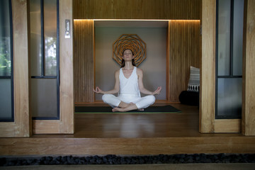 Young woman meditating in lotus position in cozy stylish studio. Woman in complitly white outfit practising yoga. Healthy lifestyle, wellbeing concept.